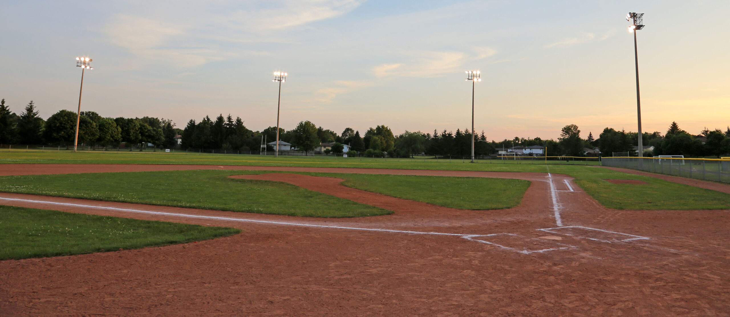 A Little League field in late summer, early evening, with the lights on.